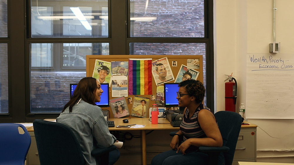 foster care education project: 2 young women smile and talk, turned toward each other in chairs at desks with computers; bulletin board with magazine covers behind them