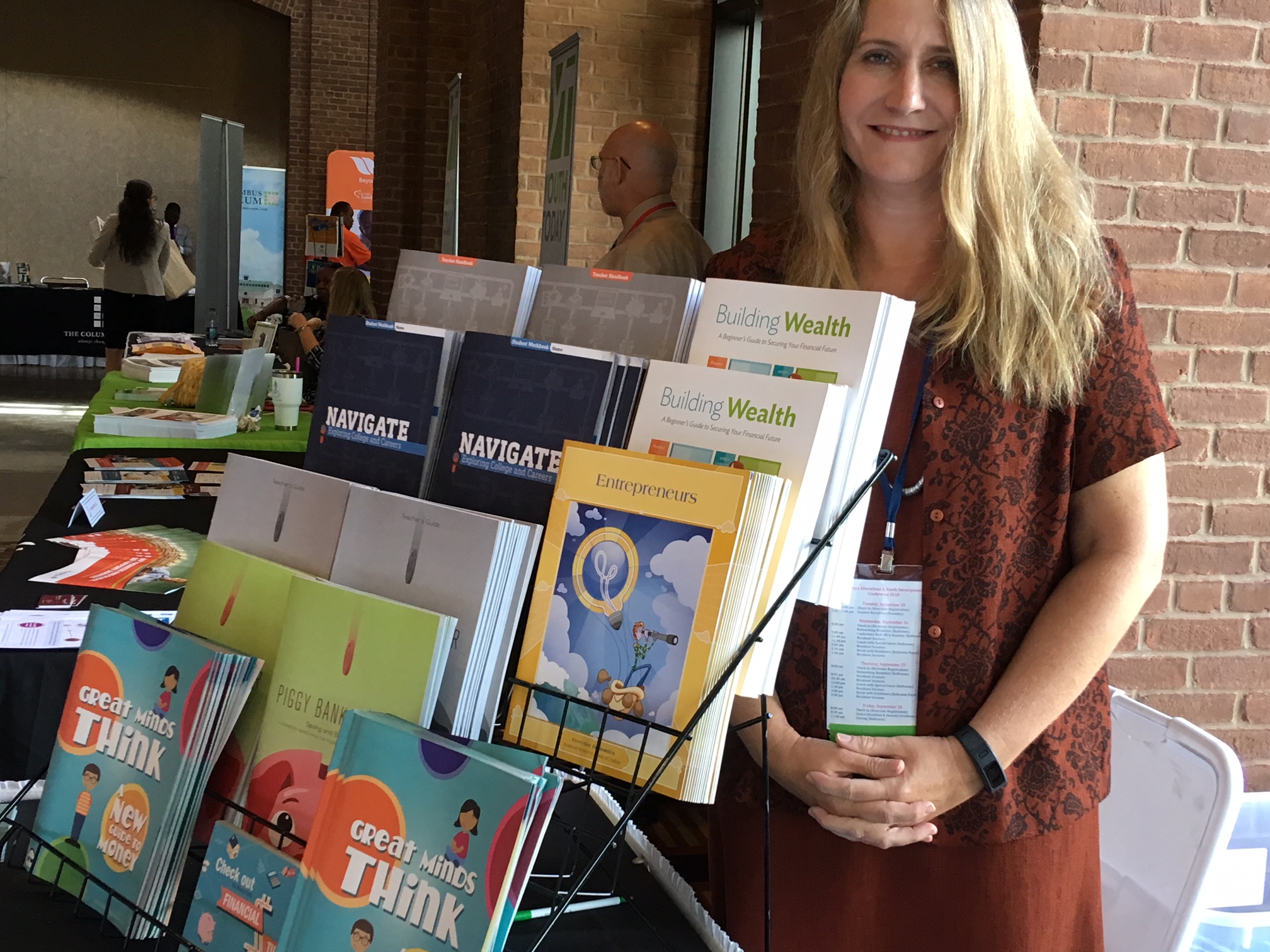 Federal Reserve Bank: Woman with long blonde hair, orange skirt and blouse stands next to table stand full of books, pamphlet about finance.