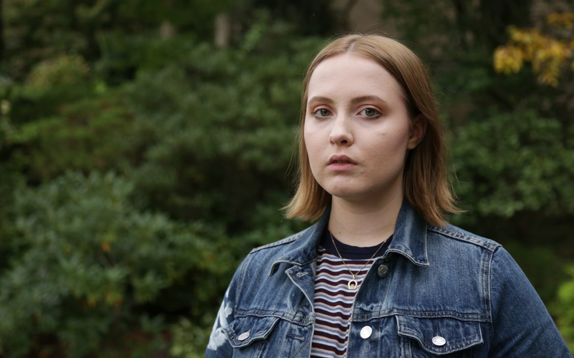 TAG: Young woman in jean jacket, striped top, necklace stares into camera