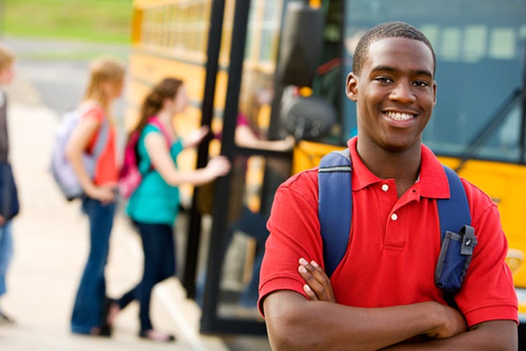 youth violence: Wholesome-looking young man wearing red polo shirt, blue backpack stands in front of schoolbus