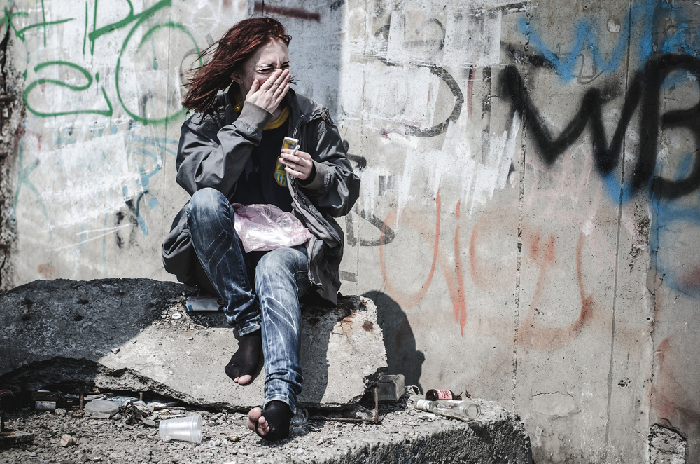 homelessness: young girl in old dirty ragged clothes, no shoes sitting on a rock, and debris scattered around holding a cigarette and a match.