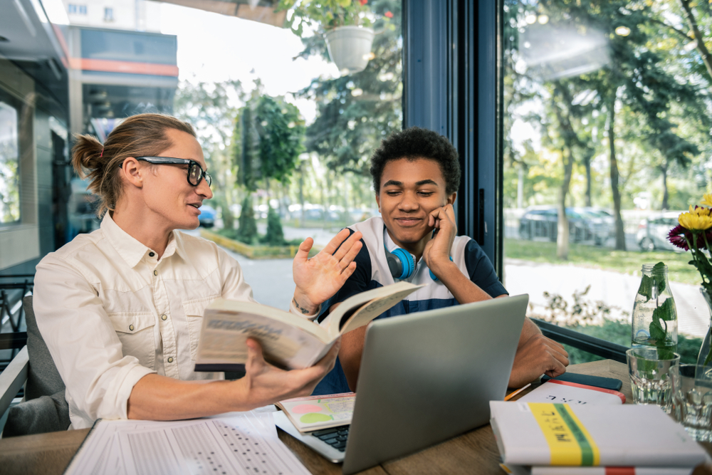 after-school: Smart young tutor holding a big book while explaining the material to the student at laptop. Outside visible through floor-to-ceiling windows.