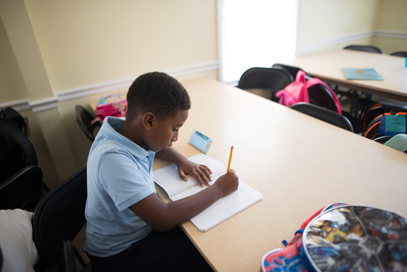 Clarkston: Boy writing in notebook at table surrounded by backpacks