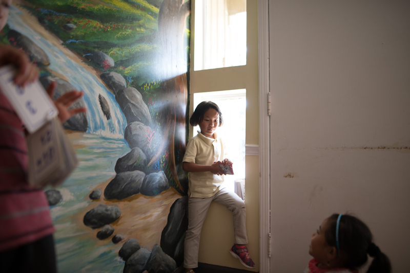 Clarkston: Little girl in yellow polo shirt, khakis sits on window sill next to mural of waterfall.