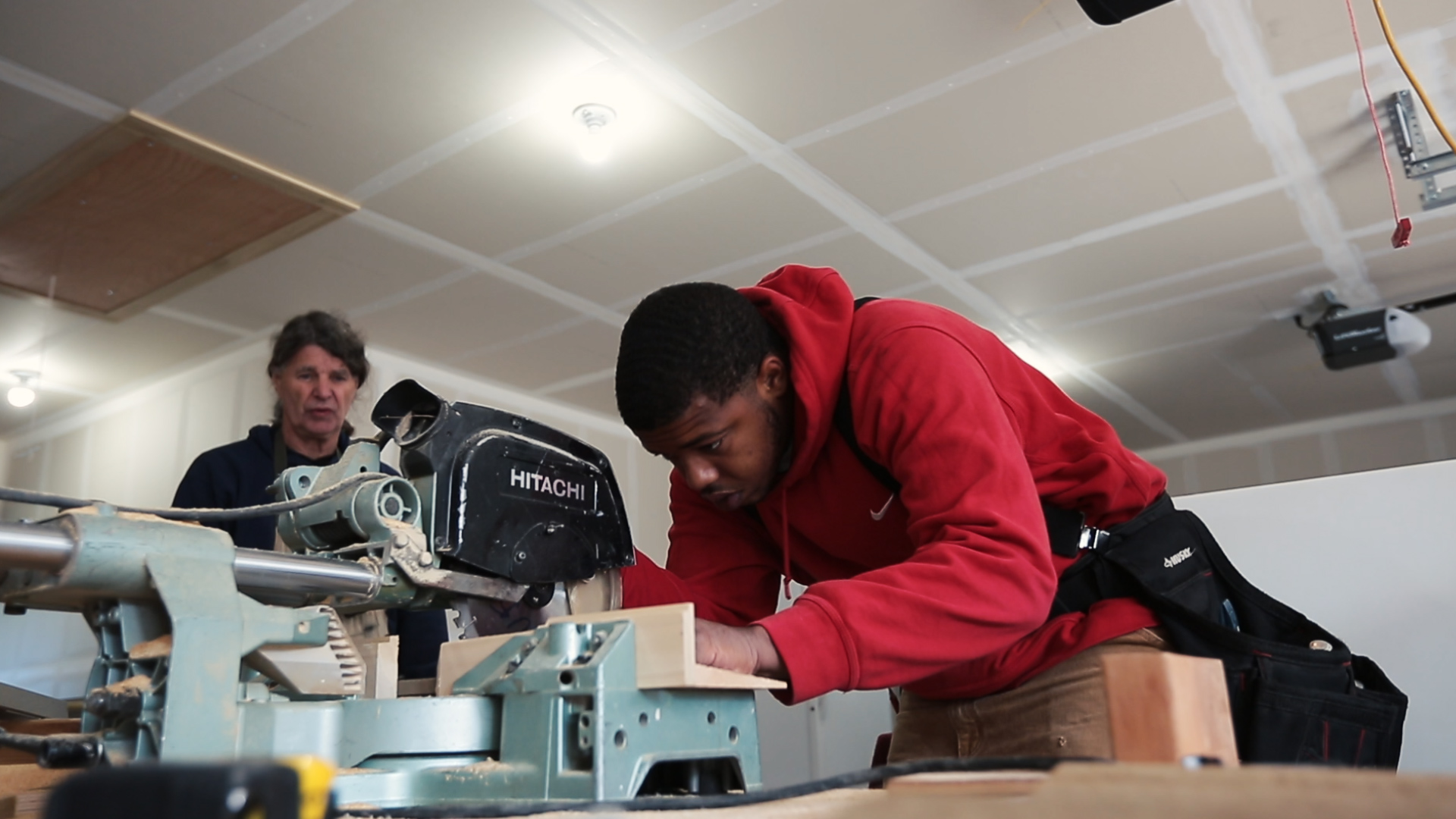 foster care: Young man in red hoodie leans over machinery, older man watches in background.