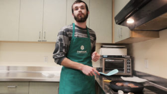 foster care: Young man with beard, mustache, wearing green apron gestures holding spatula, standing over frying pan on stove.