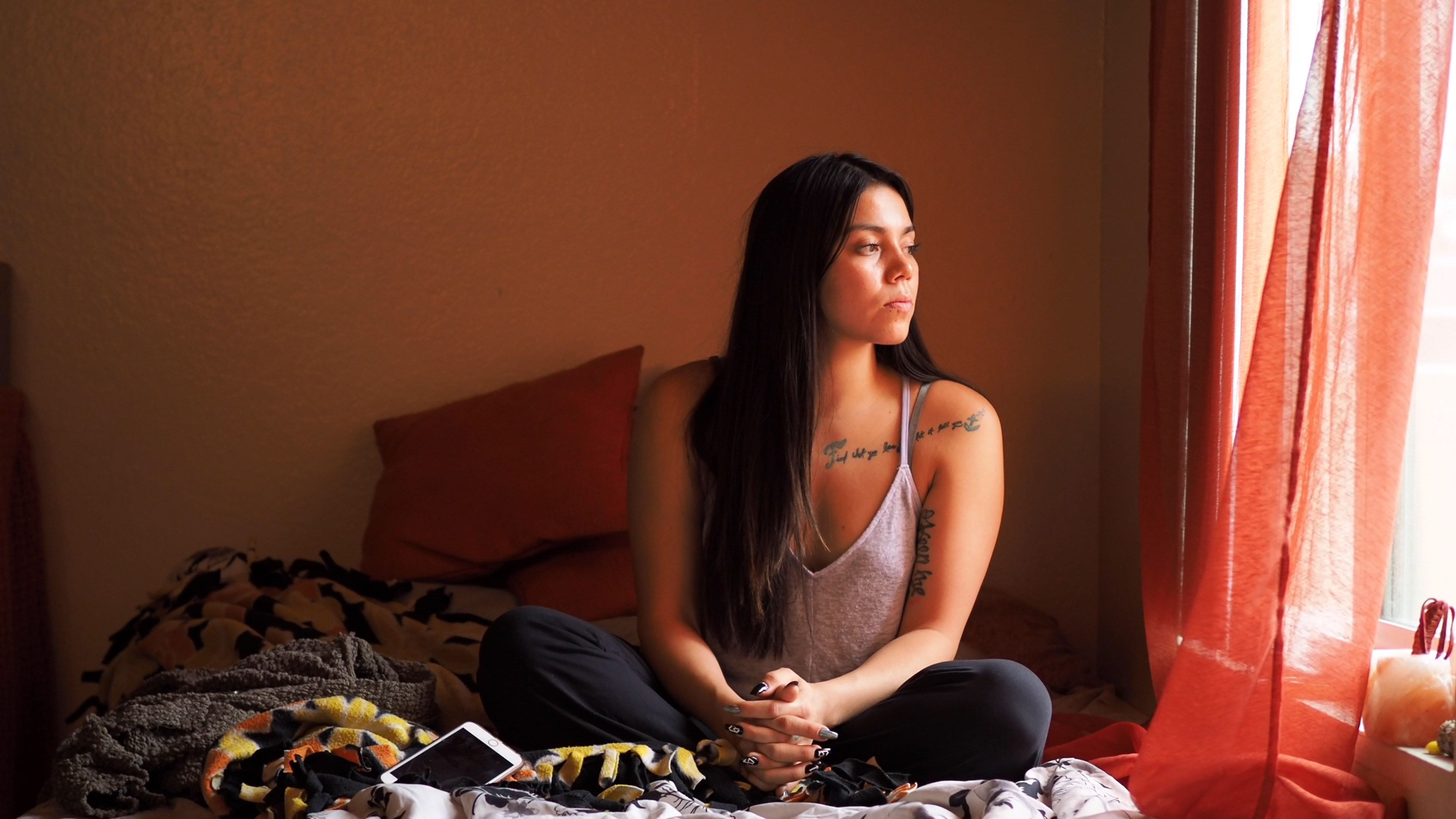 California: Young woman with long dark hair in tank top sits on bed, looks out window with orange curtains