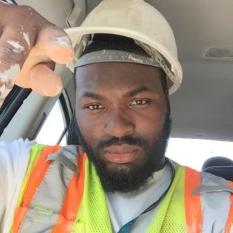 homelessness and justice project: bearded man wearing white hardhat, safety vest with white paint on fingers. 