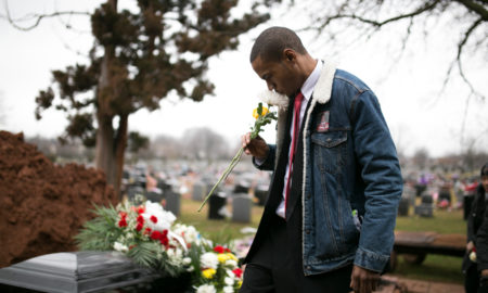 police shootings: Man in denim jacket over dark suit sniffs flower next to casket covered with flowers.