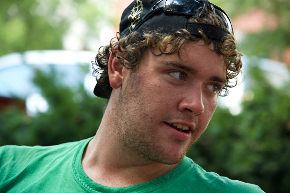homelessness and justice project: Close-up of man with stubble, blond curls, blue baseball cap worn backwards, green T-shirt.