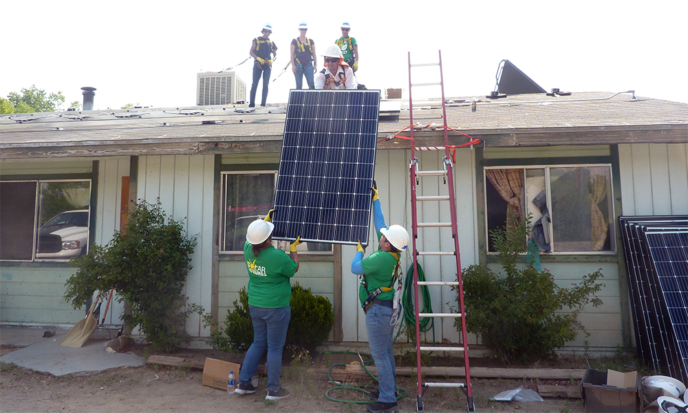 solar energy: 2 people in green T-shirts, jeans hand panel to 3 people on roof.