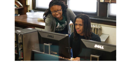 College Possible: Smiling woman with glasses bends over computer screen of young man seated at table.