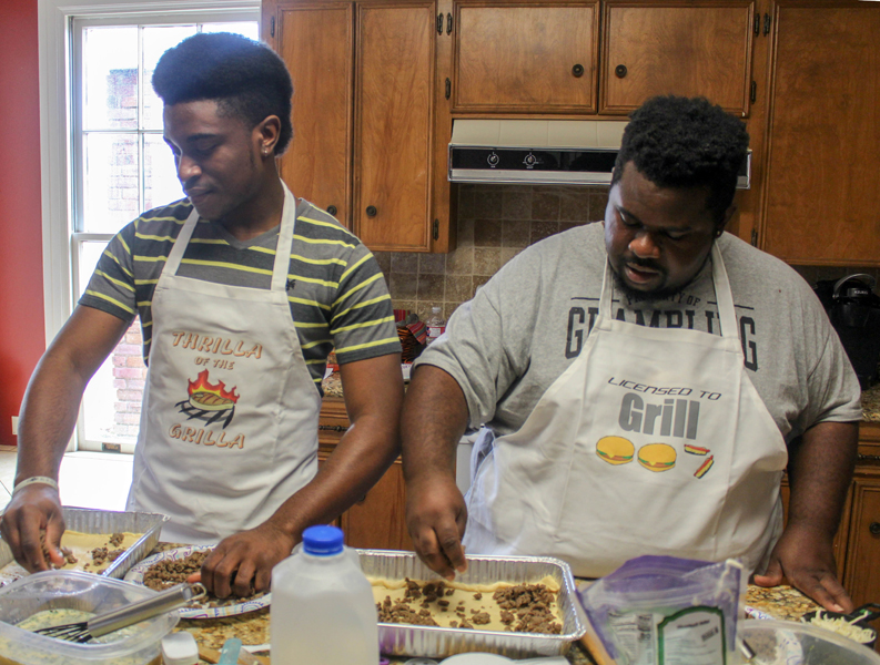 foster care: 2 young men wearing aprons assemble food at kitchen counter.