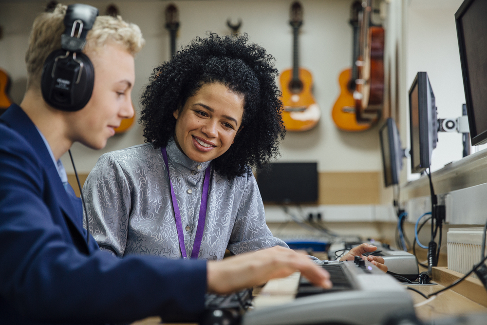 after-school: Female teacher is sitting with one of her students in a music lesson at school. He is learning to play the keyboard and is wearing headphones. 