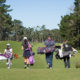 4 young people carrying golf bags, seen from back, walk through grass.