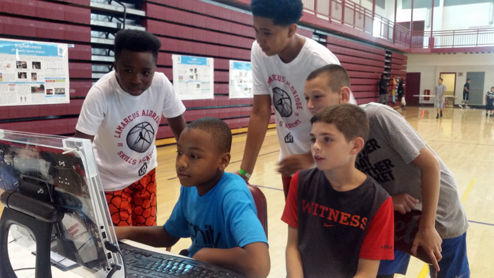 STEM: Kids in T-shirts, shorts cluster around computer on basketball court.