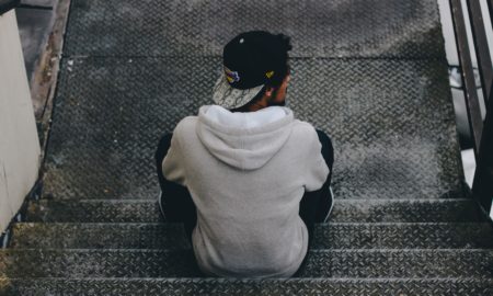 Rear view of man sitting on metal steps, wearing gray hoodie and backwards ball cap.