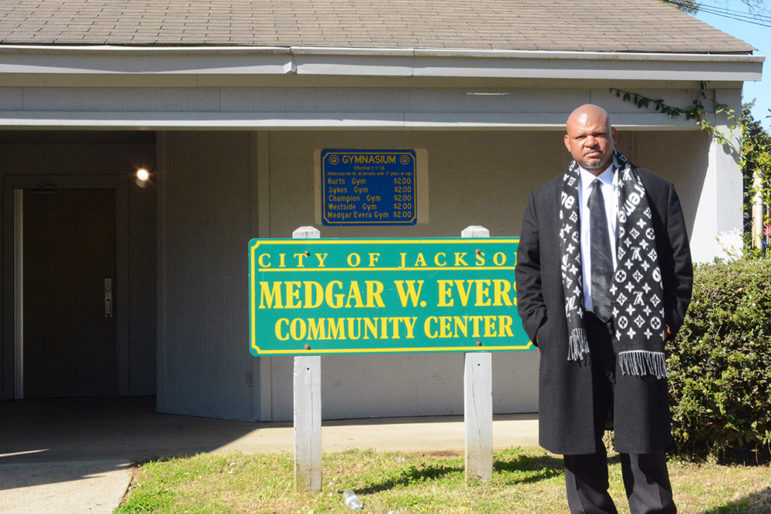 trauma: Somber man in gray coat, long patterned scarf stands in front of sign that says City of Jackson Medgar W. Evers Community Center.