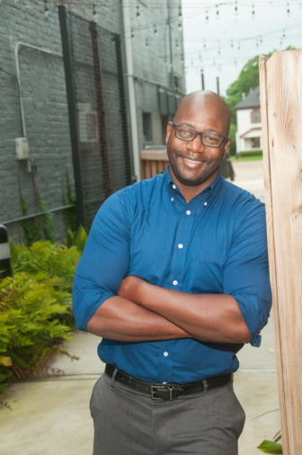 Mississippi: Smiling man with glasses, stubble wearing rolled-up blue shirt, gray pants leans against fence in alley.