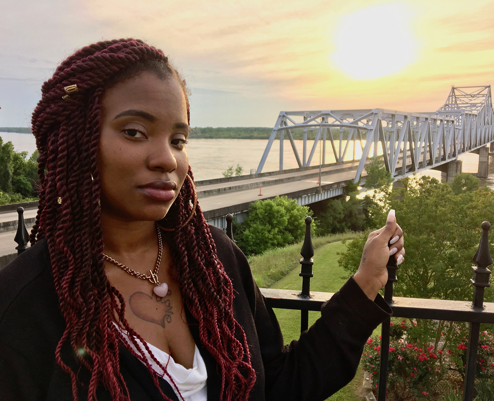 Mississippi: Somber-looking woman with long maroon braids, necklace with heart-shaped pendant holds fence post in front of bridge over river.