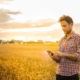 4-H: Man in plaid shirt working on tablet in front of wheat field.