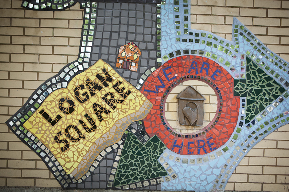 Mosaic on yellow brick wall, wth words “Logan Square” and “We are here,” within the mosaic alone with a small house with a heart in the doorway.