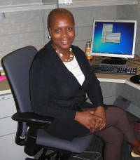 Smiling woman in suit, necklace, earrings, sitting at desk in office.