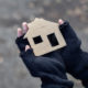 Partially gloved mittens holding a cardboard cutout of a house, with fuzzy background.