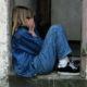 Young girl covering her face with her hands, sitting in a doorway, wearing jeans and a jean jacket and sneakers.