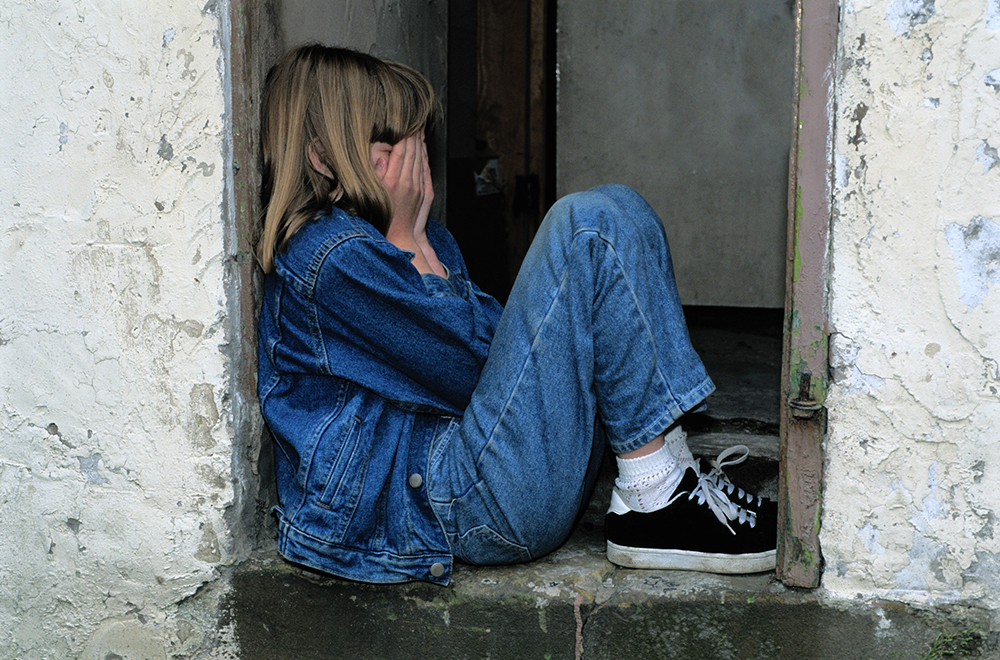 Young girl covering her face with her hands, sitting in a doorway, wearing jeans and a jean jacket and sneakers.