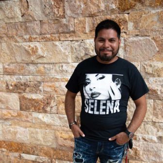 New Mexico: Smiling man with beard, mustache, wearing black Selena T-shirt, distressed jeans, stands in front of wall.