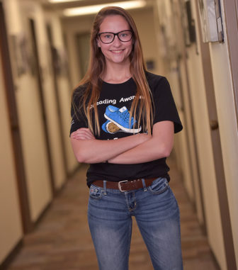 campus coach: Smiling young woman with long brown hair, glasses, T-shirt, jeans stands in hall with arms folded.