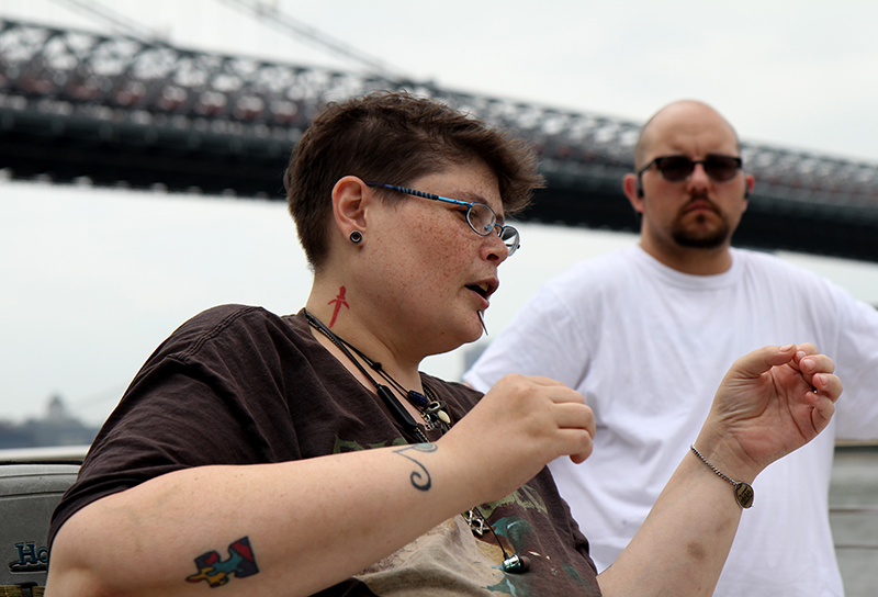 LGBTQ: Person in T-shirt with short brown hair, glasses, earlobe plugs, necklaces, tattoos, spike above chin gestures, looking to right. Balding man in background with beard, mustached, white T-shirt, sunglasses looks on.