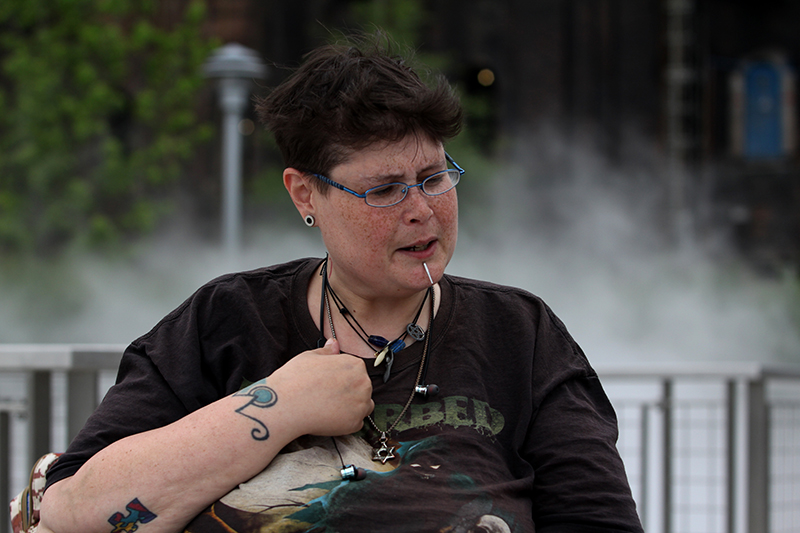 LGBTQ: Person in T-shirt with short brown hair, glasses, earlobe plugs, necklaces, tattoos, spike above chin looks down.