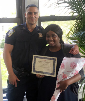 NYPD: Police officer smiles with arm around shoulder of smiling young woman holding up certificate, flowers.