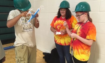 drones: 3 young people wearing green hard hats hold drone, controller.