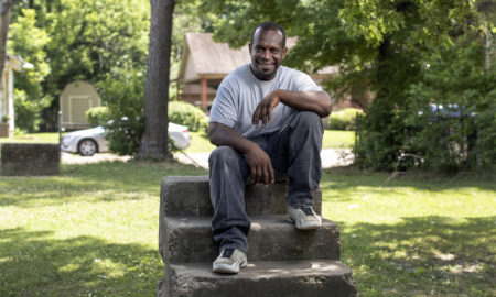 gang violence: Smiling man in T-shirt, jeans, sneakers sits on top of cement short set of stairs on lawn.