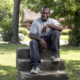 gang violence: Smiling man in T-shirt, jeans, sneakers sits on top of cement short set of stairs on lawn.