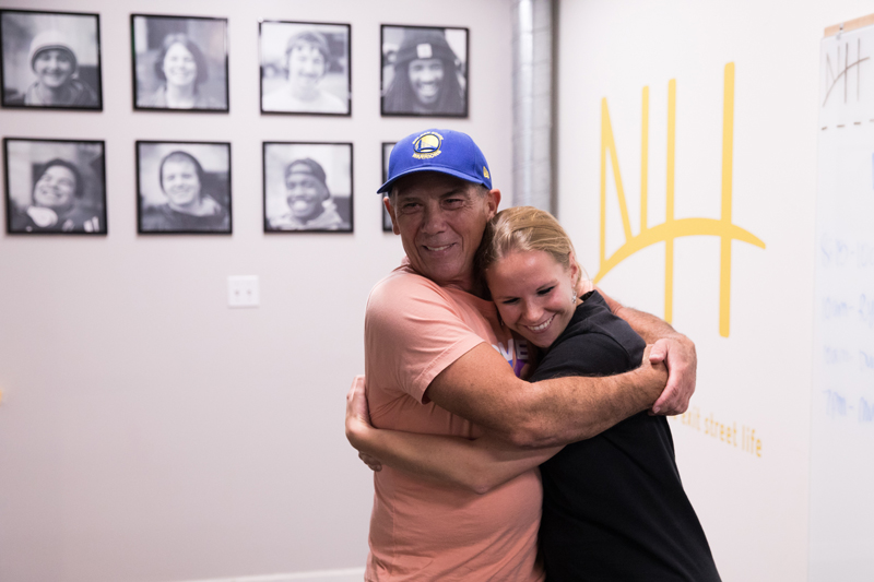Homelessness: Smiling man and woman hug in front of wall of photos.