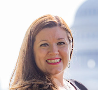 Homeless: Darla Bardine (headshot), executive director of the National Network for Youth, smiling woman with long brown hair wearing black sleeveless dress, necklace, earrings standing in front of U.S. Congress