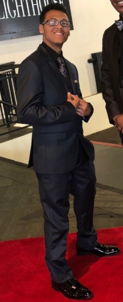 Housing First: Lyndon Hernandez (headshot), youth peer advocate/community organizer, smiling man with glasses, dark suit, dark shirt, dark tie, dark shoes 