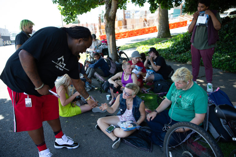 Tacoma: Man in dark blue T-shirt, bright red shorts hands something to young people sitting on ground.