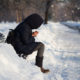 youth homelessness: lonely person with hoodie and backpack staying on bench in city park with snow