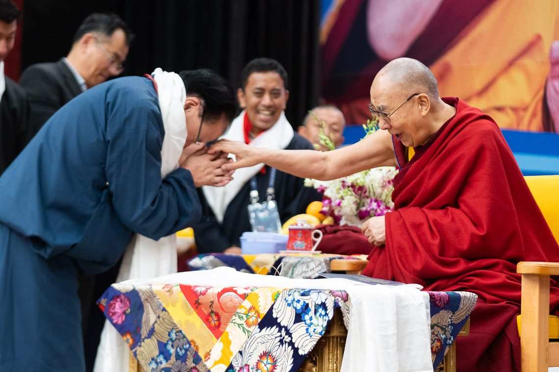 Dalai Lama: Standing man in blue bows forehead to hand of man seated at table in red robe, glasses, shaved head
