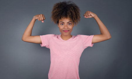 foster care: Waist up shot of African American woman raises arms to show her muscles feels confident in victory, looks strong and independent, smiles positively at camera, stands against gray background.