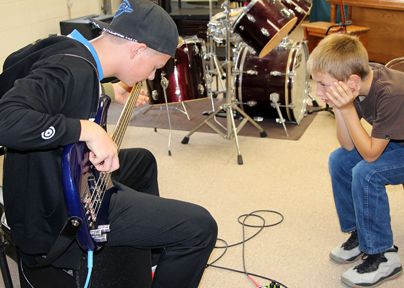 four-day school week: Boy at left plays guitar while another watches him, both seated near drum setup.