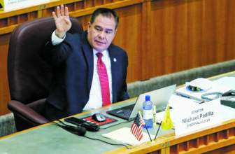 New Mexico: Man sitting behind long desk raises left hand, looks to left. Name card on desk says senator Michael Padilla.
