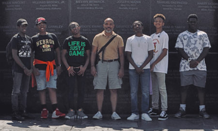 black boys: 6 boys in T-shirts and jeans with smiling man in middle wearing polo shirt and shorts in front of memorial.
