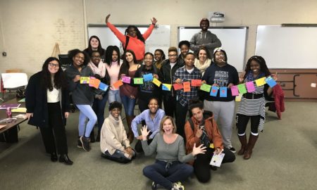 girls: Big group of smiling young women pose with cards on a string; smiling woman in gray sweater, jeans, sneakers poses in front of them with jazz hands