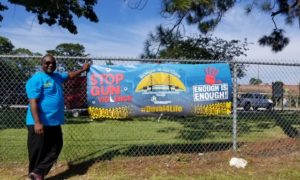 Paul Tutwiler: Man in sunglasses, blue T-shirt, black pants, brown shoes stands to left of poster on fence that says stop the gun violence Duval4life enough is enough